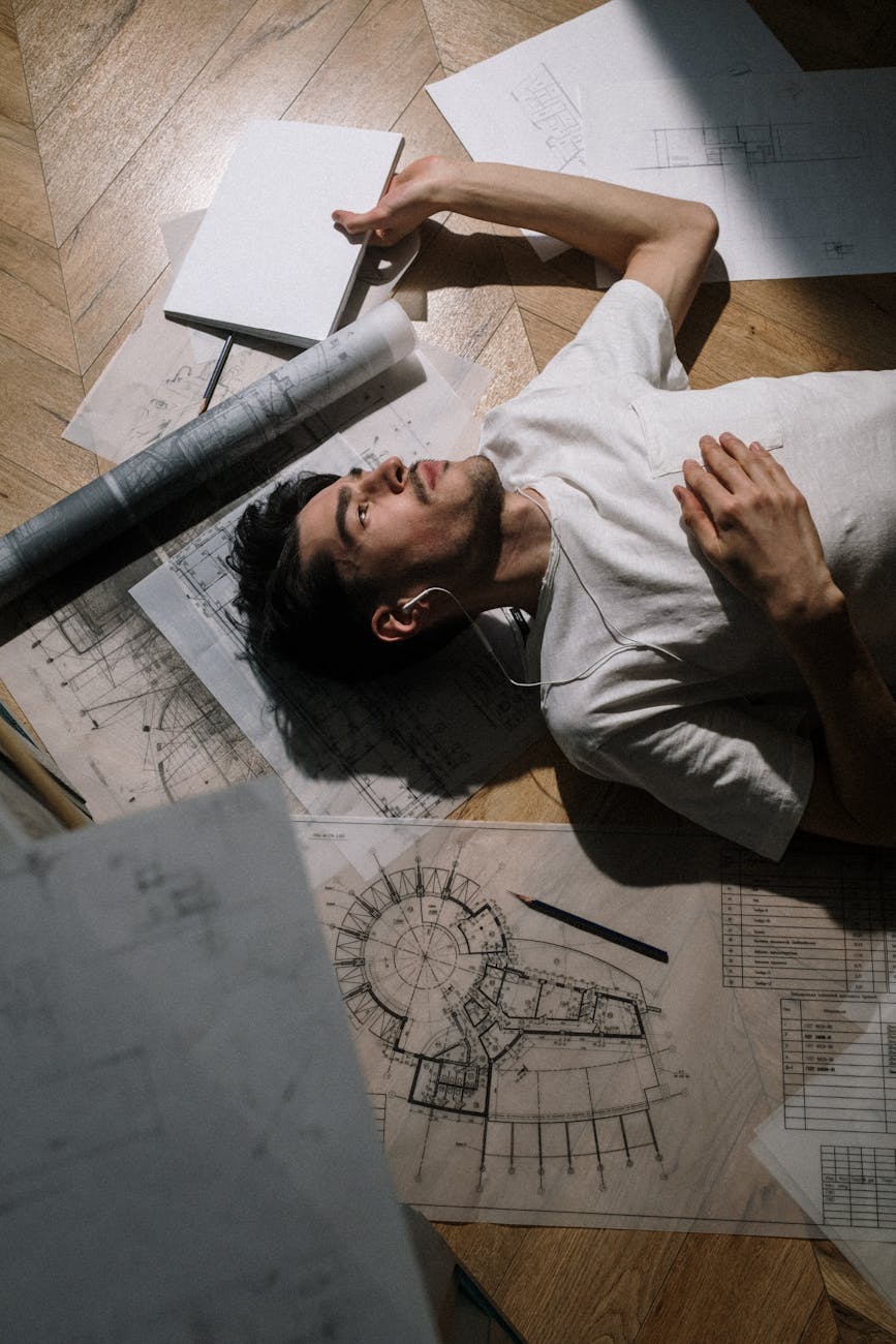 An architect with earphones lies on floor surrounded by architectural drawings on a wooden floor.