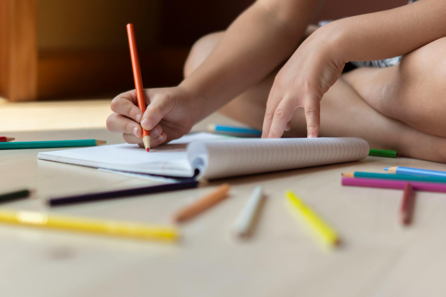 Ground level of unrecognizable kid sitting on floors with legs crossed and drawing in copybook with multicolored pencils
