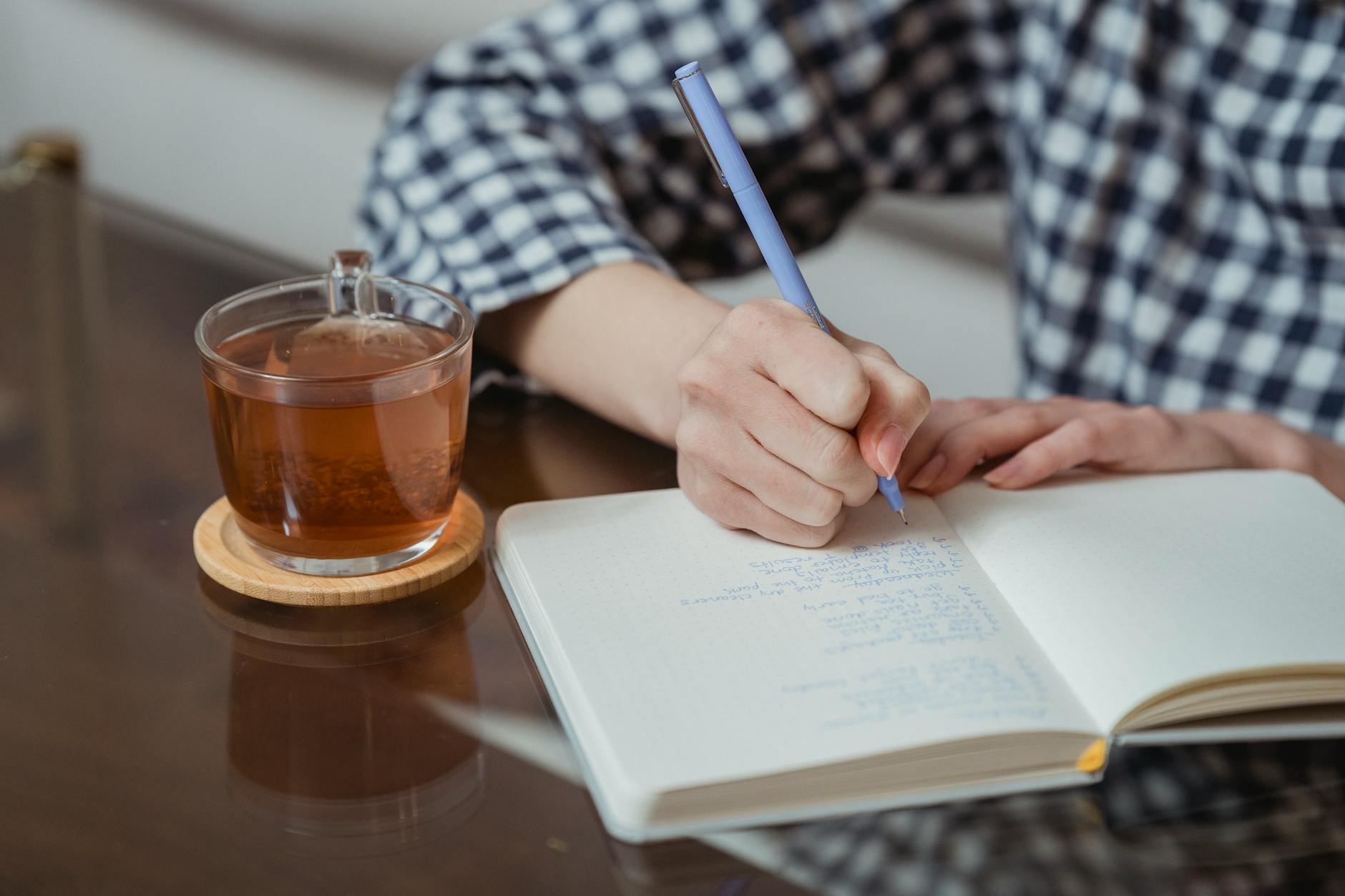 Close-up of hands writing in a notebook beside a glass of tea on a wooden coaster.