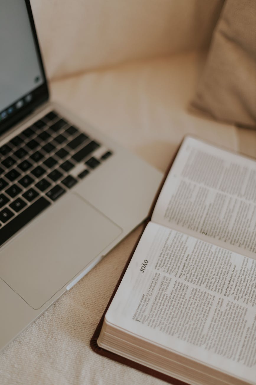 An open Bible next to a laptop on a cozy bedspread, ideal for studying or devotional moments.