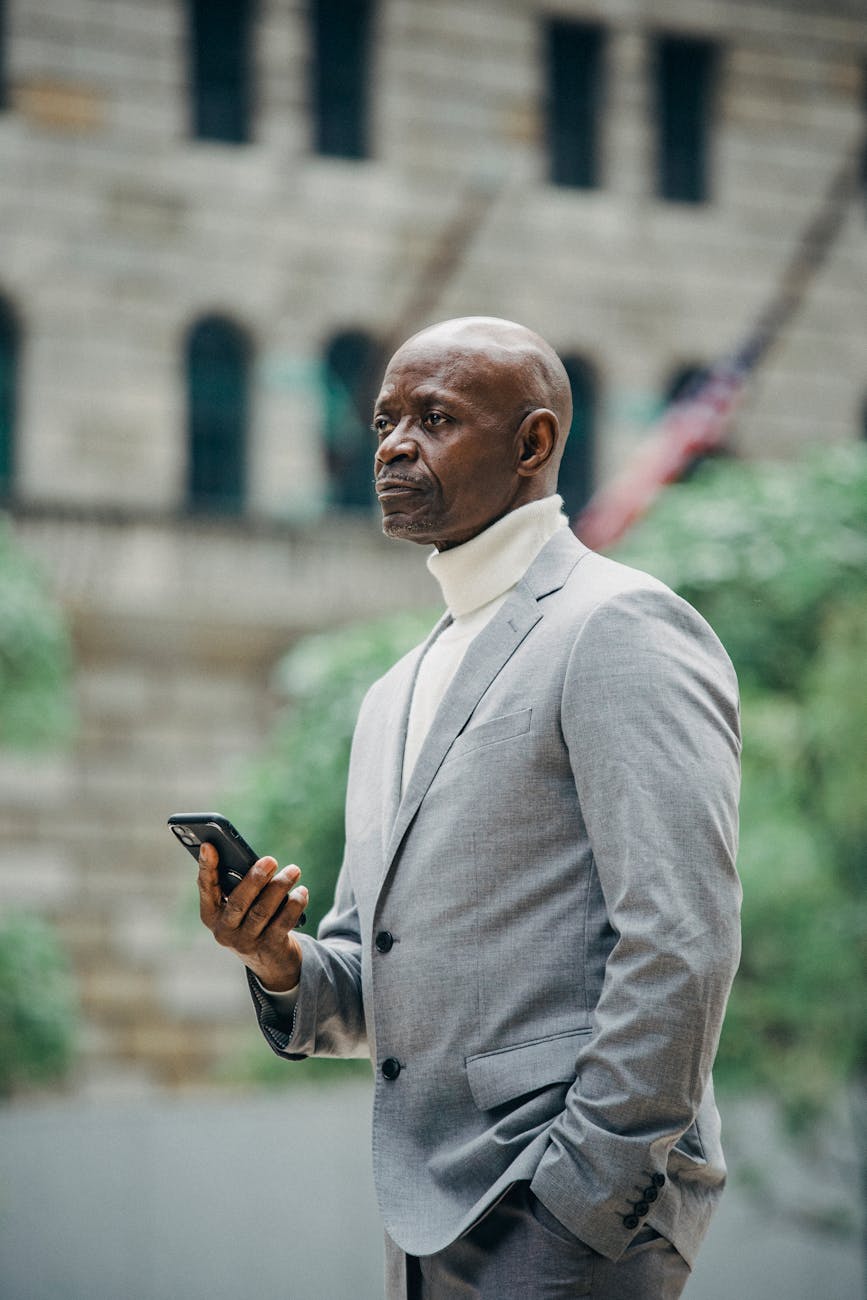 Elegant African American man in a suit holding a smartphone, standing outdoors in a downtown area.