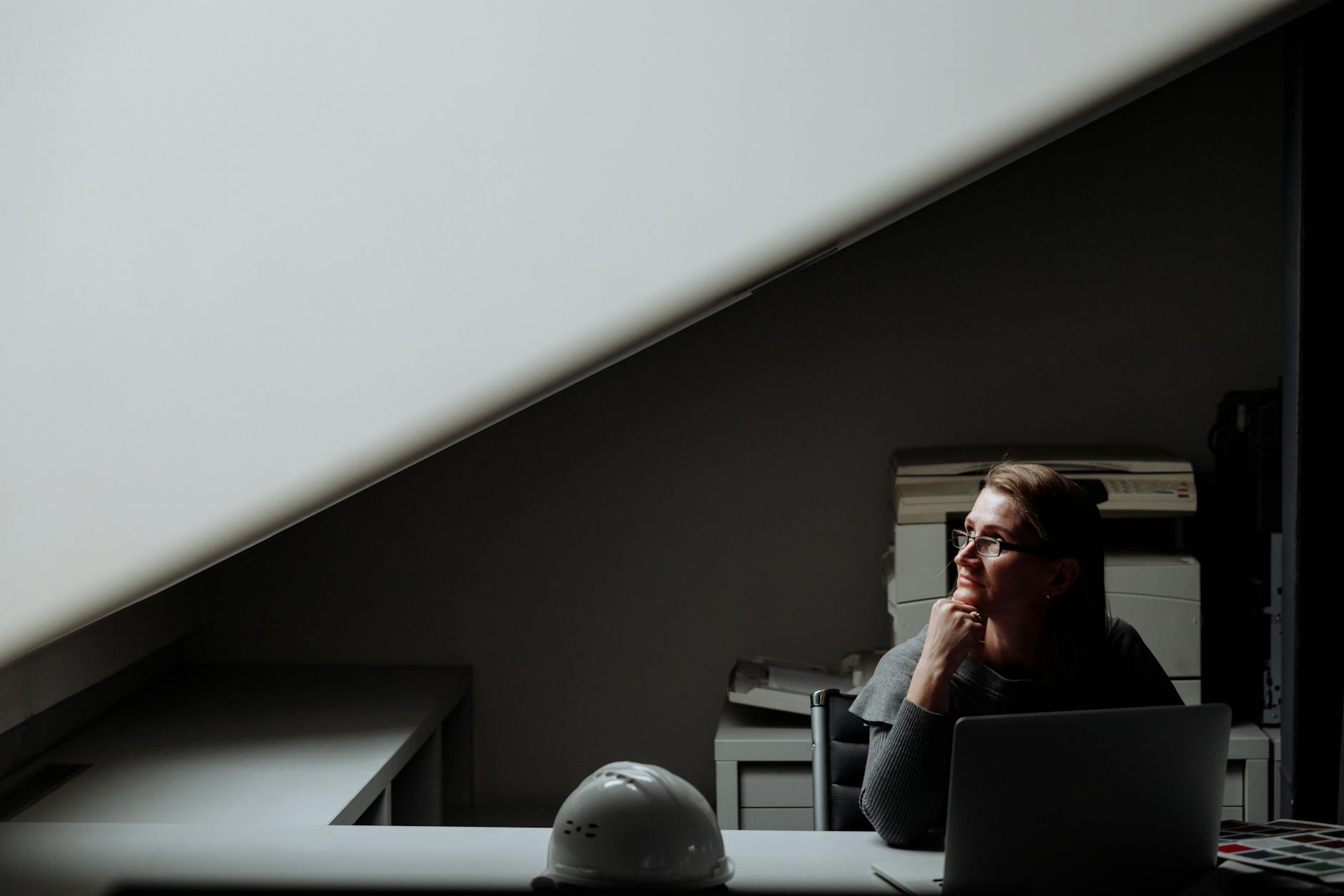 A woman sits at a desk in a dimly lit office, contemplating while using a laptop.