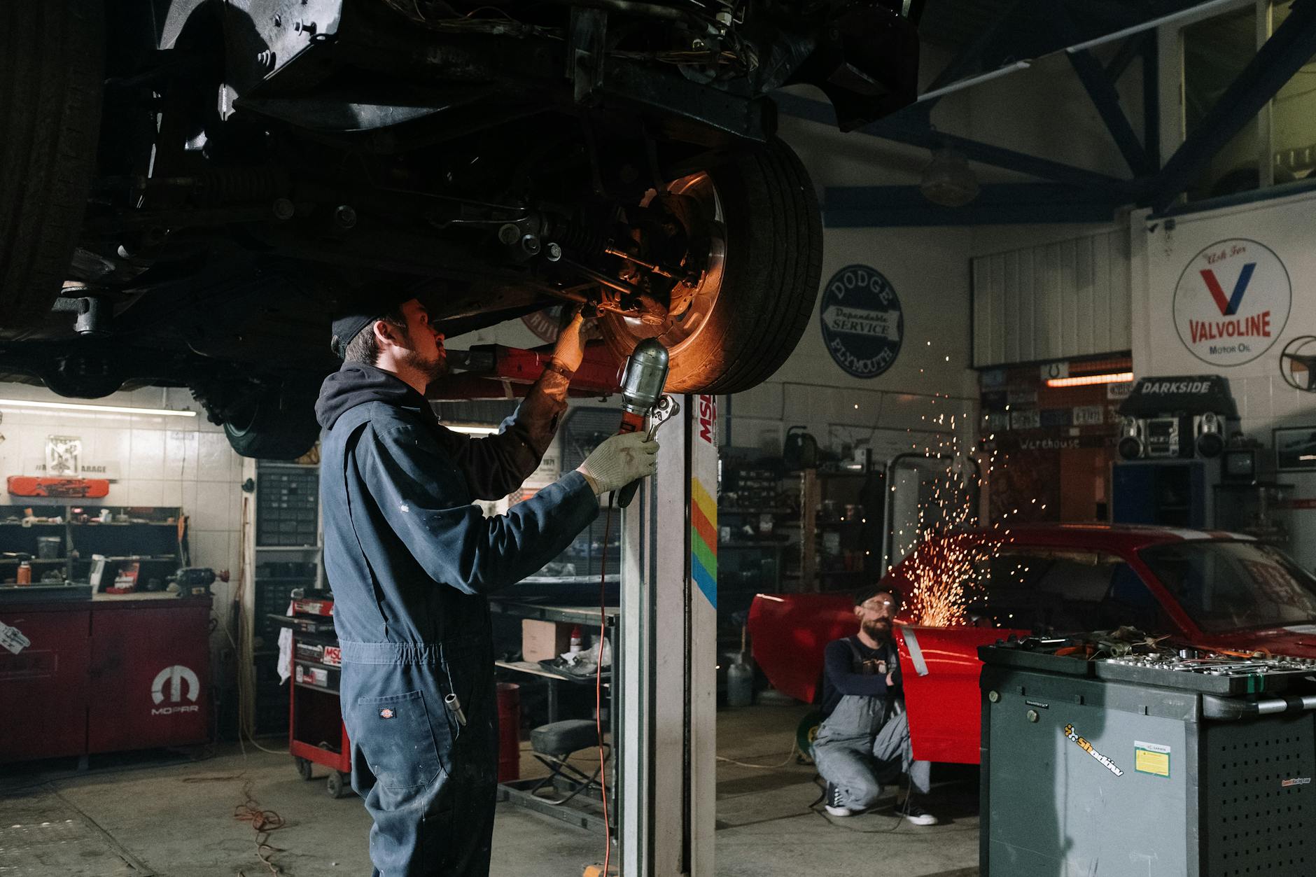 Two automotive technicians repairing a car in a busy workshop with sparks flying.