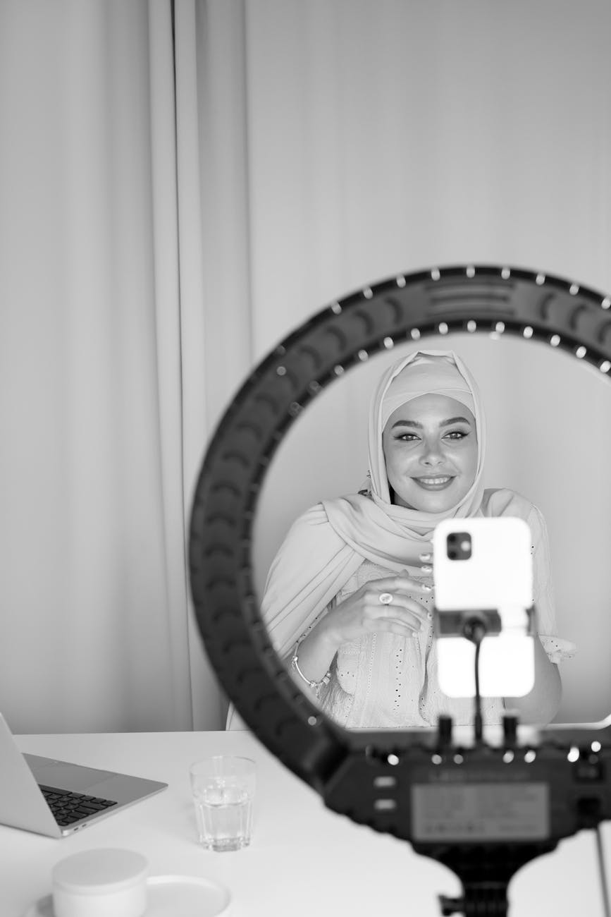 Black and white image of a Muslim woman using a ring light and smartphone for vlogging indoors.