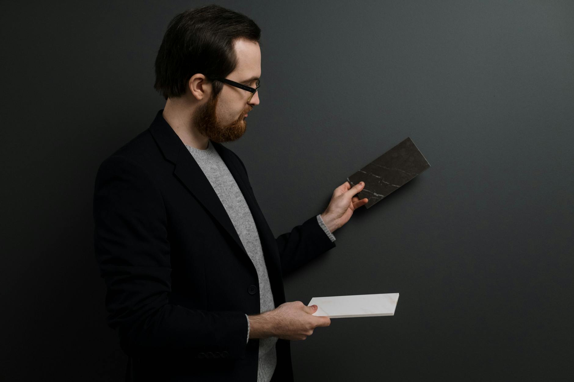 Man in a dark blazer holding and comparing two tile samples against a dark background.