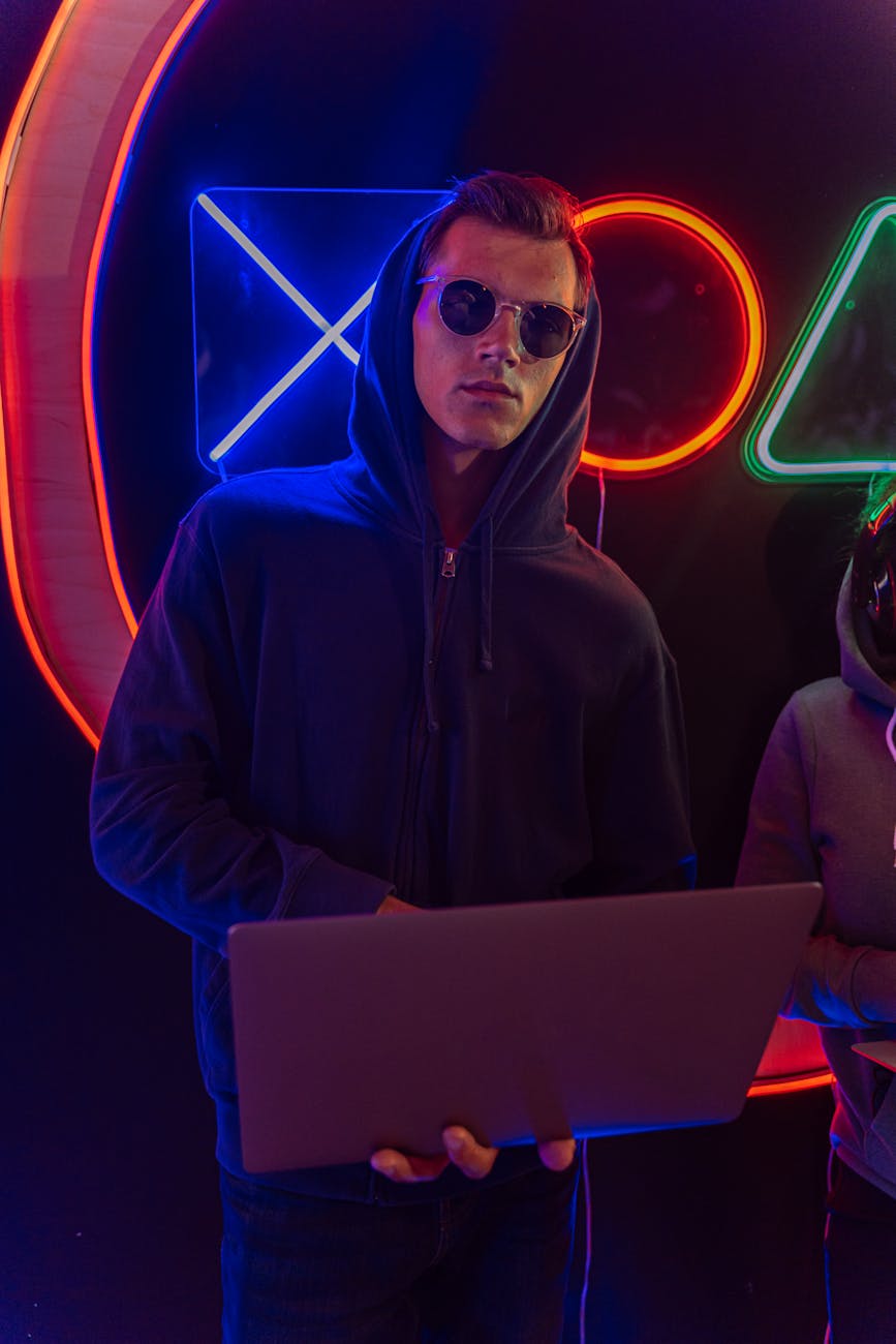 Man in hoodie with laptop in neon lights, depicting cybersecurity threat.