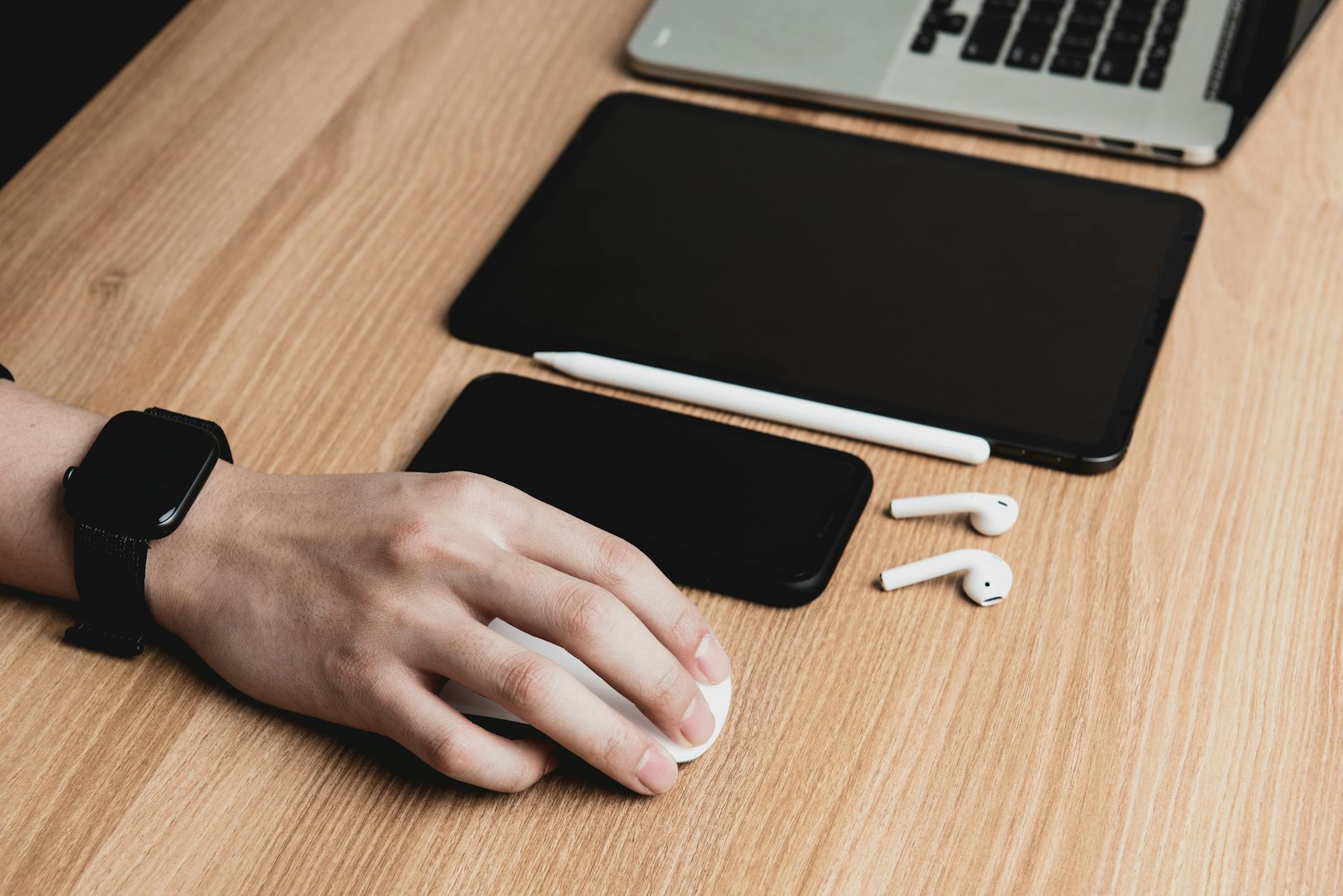 A hand using a mouse with a laptop, tablet, smartphone, and earphones on a wooden desk.