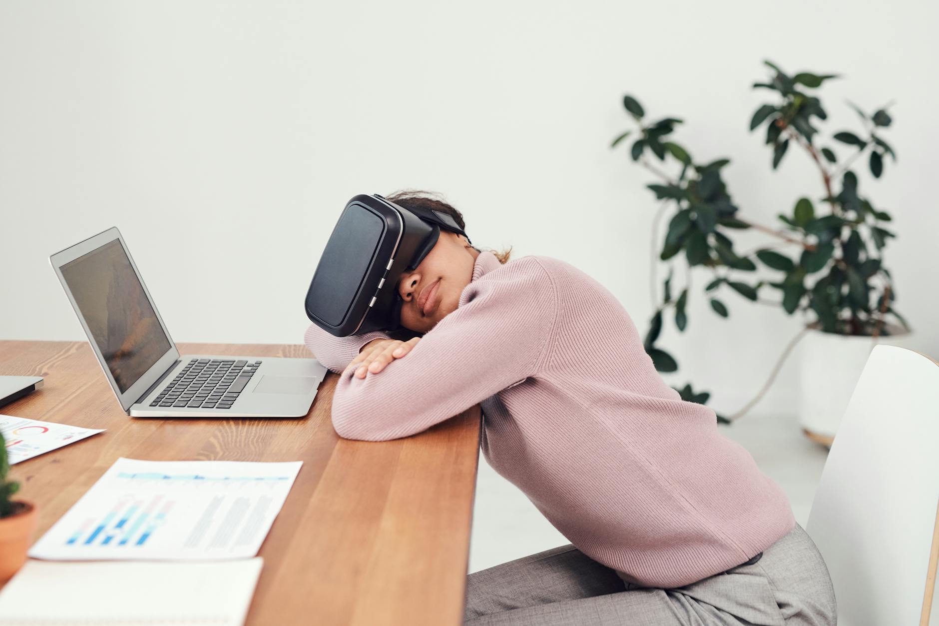 A woman wearing a VR headset rests on a desk beside a laptop, combining relaxation and technology.