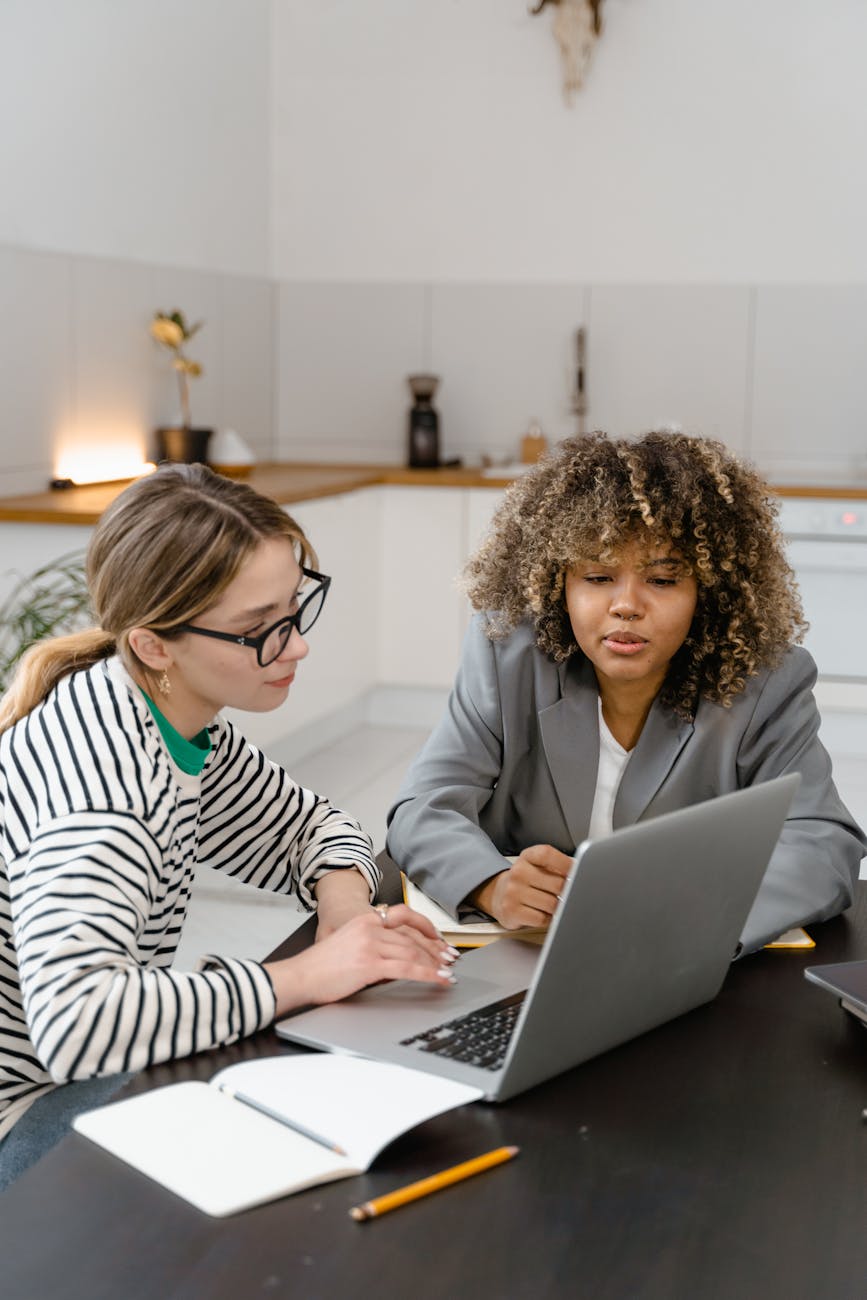 Two women discuss work strategy at a laptop in a modern office.
