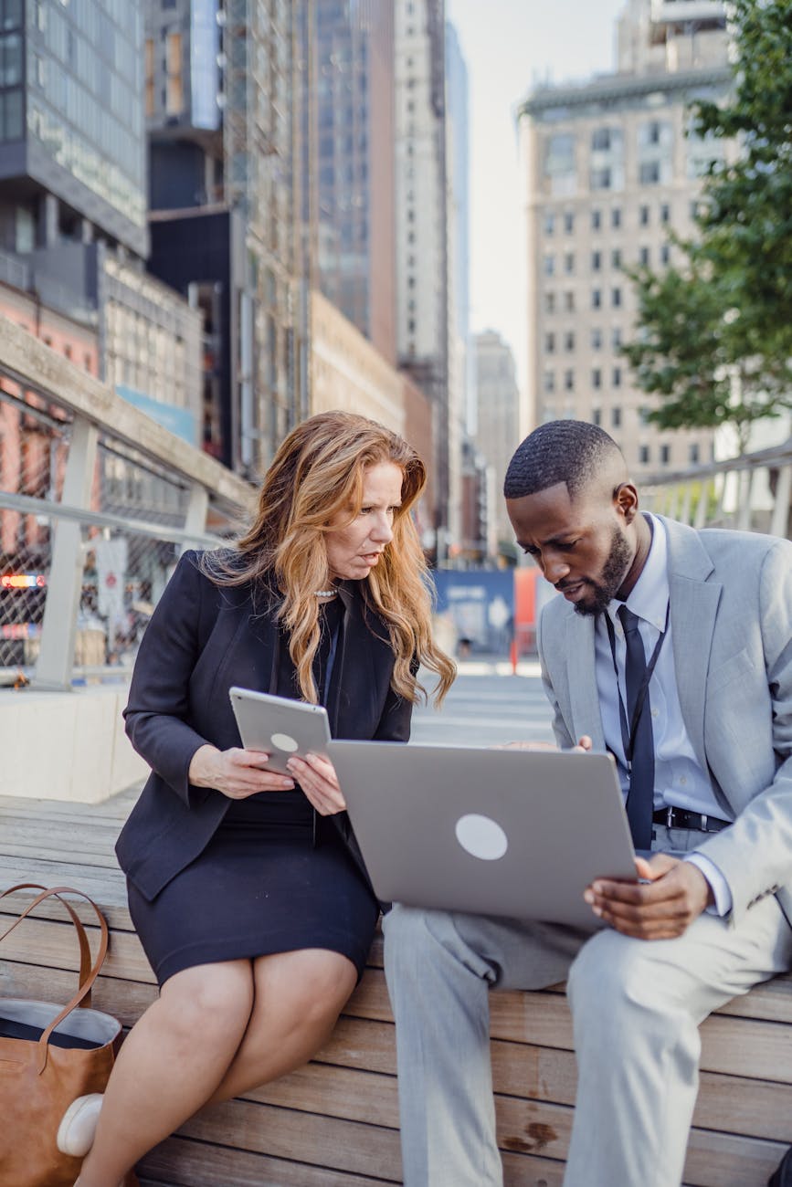 Two business professionals discuss work on a laptop and tablet outdoors in an urban setting.