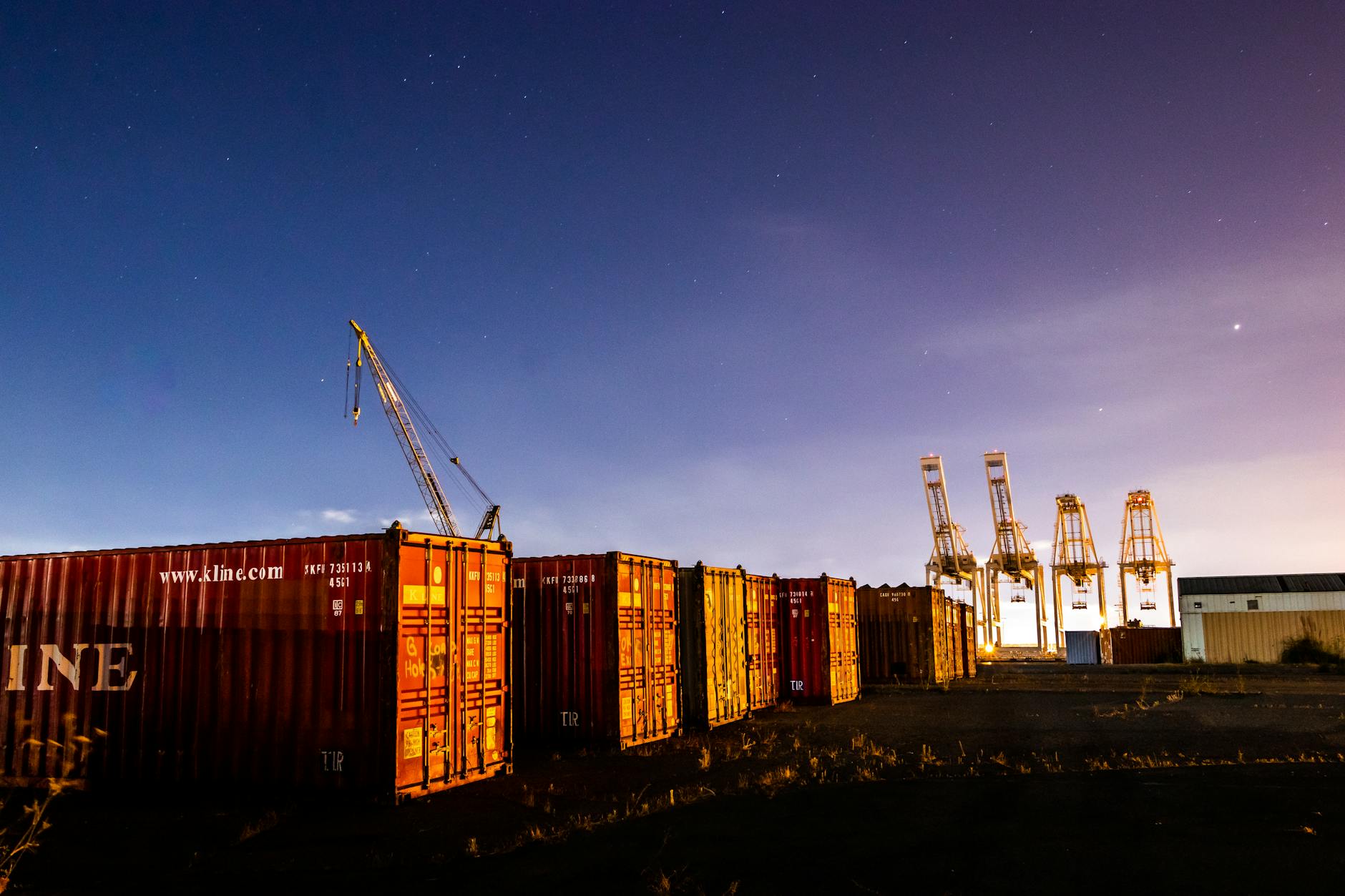 A scenic night shot of a shipping yard with cargo containers and cranes under a starry sky.
